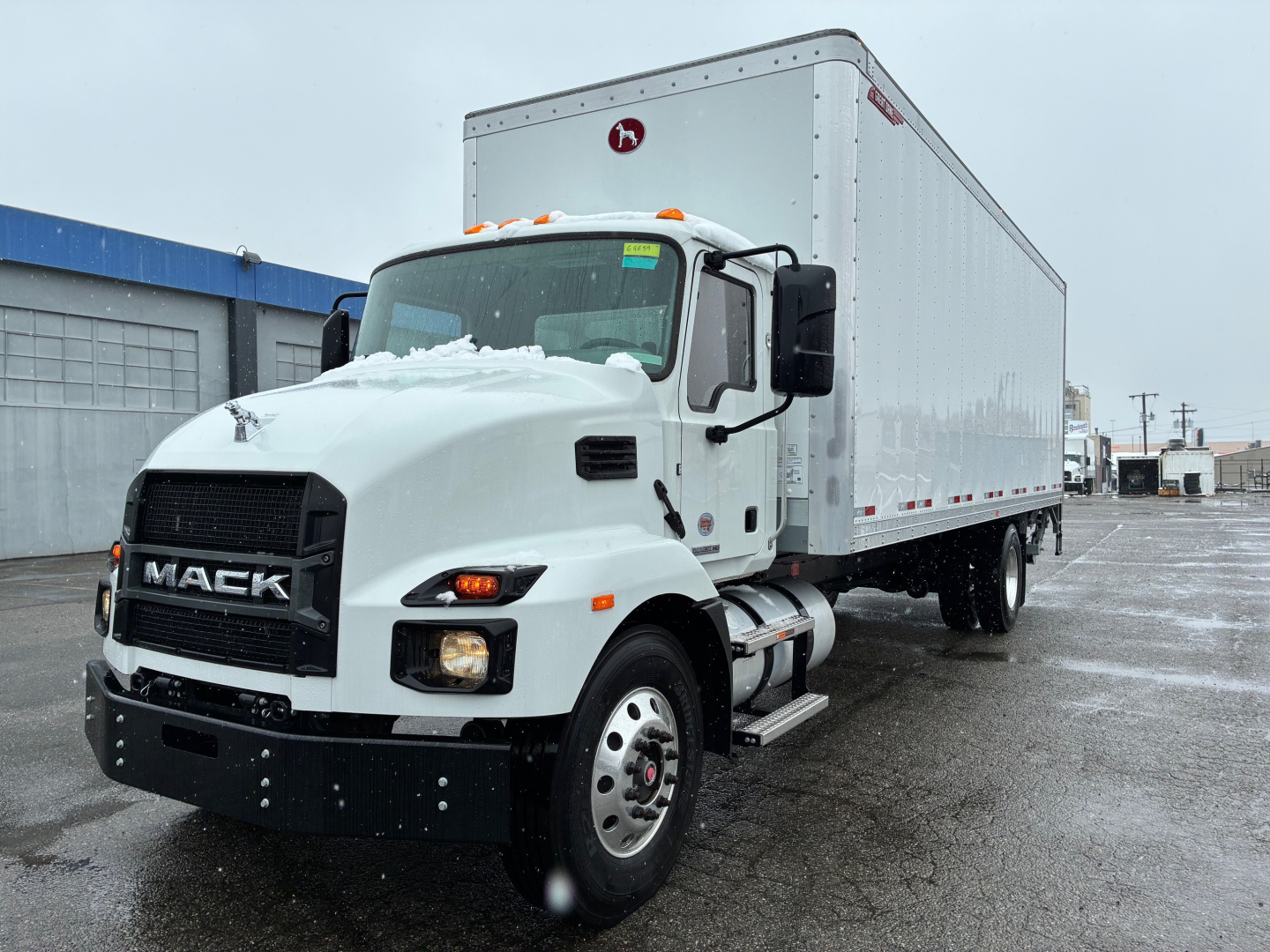 Mack MD6 box truck in white, front-left view showing cab with black grille, large dual rear-view mirrors, and long box trailer; snow on hood and ground.
