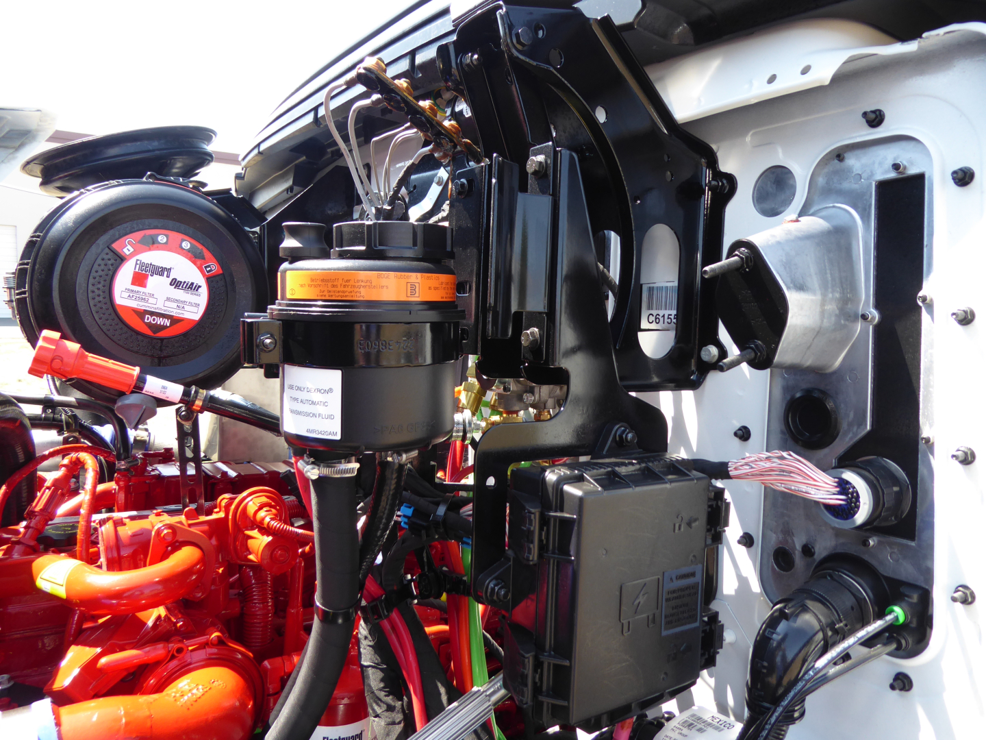 Engine bay of 2025 Mack MD6 showing orange battery/air reservoir, black electrical box, wiring harness, and front chassis components.