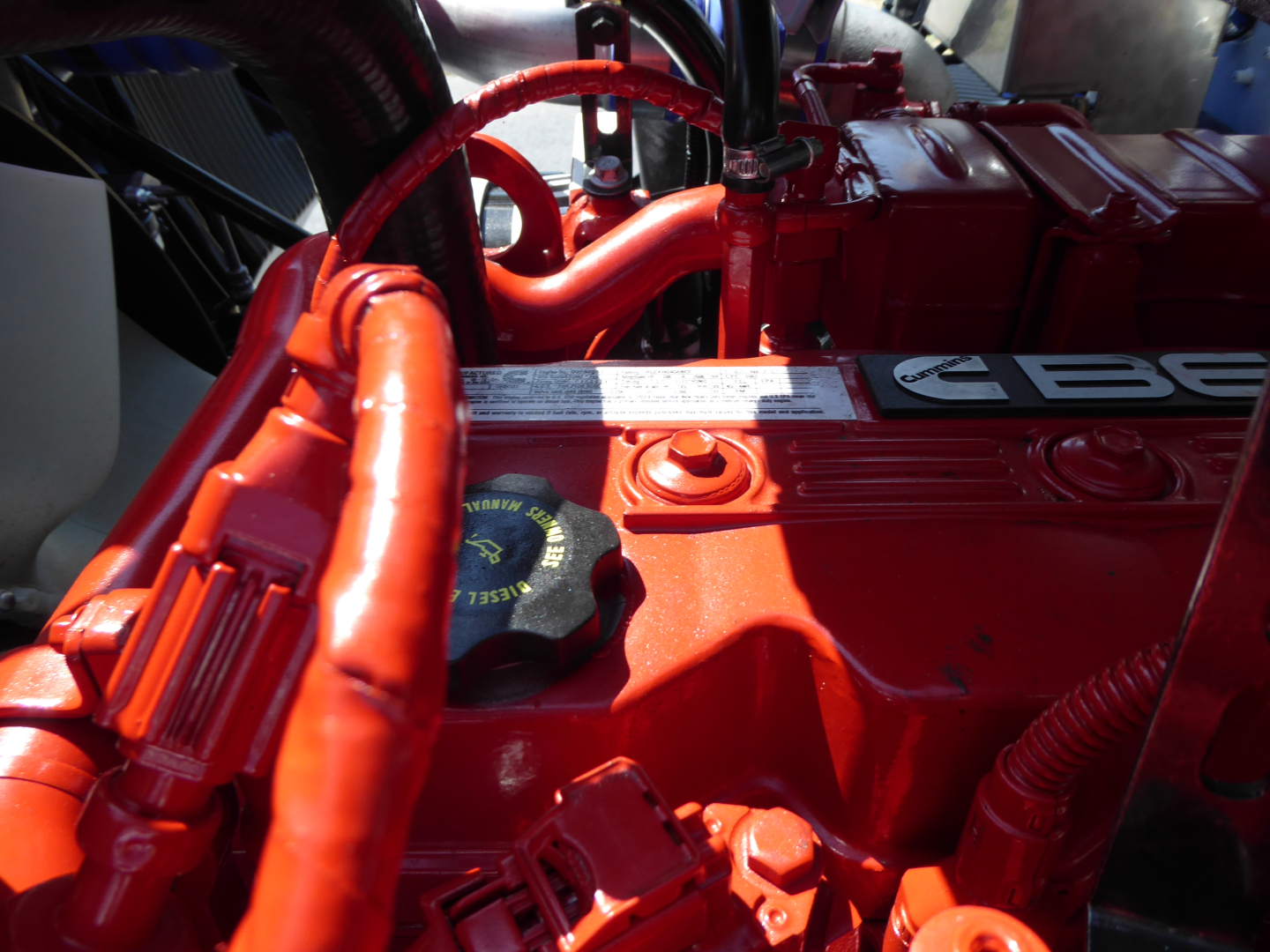 Engine bay of a 2025 Mack MD6 showing bright orange/red components, hoses, and a labeled valve cover with engine oil cap visible.