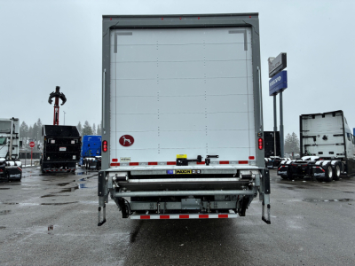 Rear view of a new 2025 Mack MD6 box truck with a rear liftgate, reflecting light with a license plate area and company logos on the door.