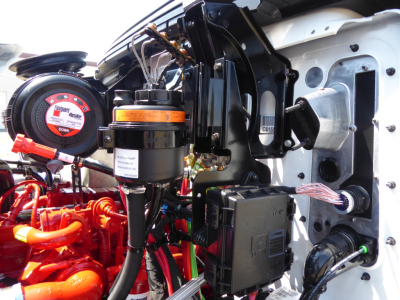Engine bay of 2025 Mack MD6 showing orange battery/air reservoir, black electrical box, wiring harness, and front chassis components.