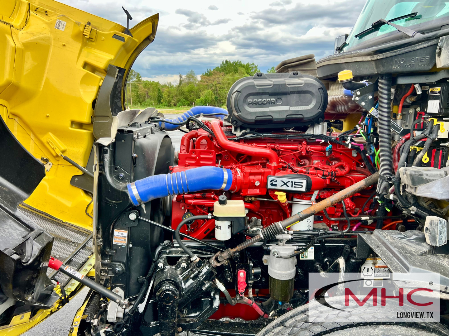 Peterbilt 579 engine bay with red Cummins ISX15 engine, blue hoses, black air intake, yellow hood, and various components visible.