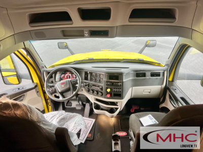 Peterbilt 579 cab interior from front passenger seat showing dashboard gauges, steering wheel, center console, and driver’s seat. Yellow exterior visible through windshield.
