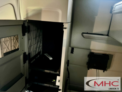 Truck cab interior with storage compartments and a small black device mounted, Peterbilt 579. Visible interior panels and a badge/logo 'MHC Longview, TX' corner.