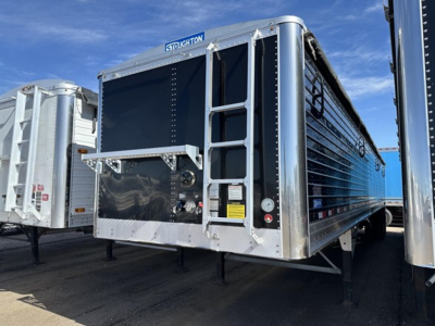 Stoughton grain trailer with hydraulic tail service doors and black interior; visible labels include 'STOUGHTON' on top front, VIN tag area, and safety stickers on side; aluminum framing and ladder rungs along the front corner; blue and sil