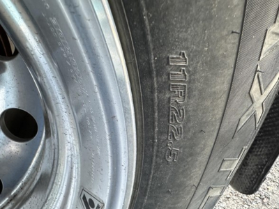 Trailer tire and wheel with embossed tire size 11R22.5/AXLE on sidewall; rim metallic with some dirt.