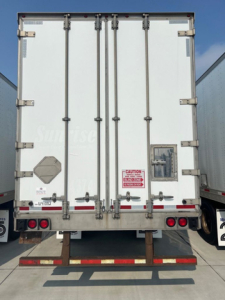 Rear view of a white utility van trailer with double doors, taillights, safety stickers, and a wooden bumper protruding from the bottom.