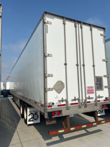 Rear view of a white utility trailer with double rear doors, reflective tape, and bumper; small red warning label and diamond placard on door; mudflaps show '25'