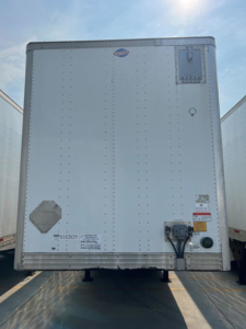 Rear view of a white utility van trailer showing jack assembly and various decals; small diamond-shaped plate, circular access port, and a rectangular panel near top right.