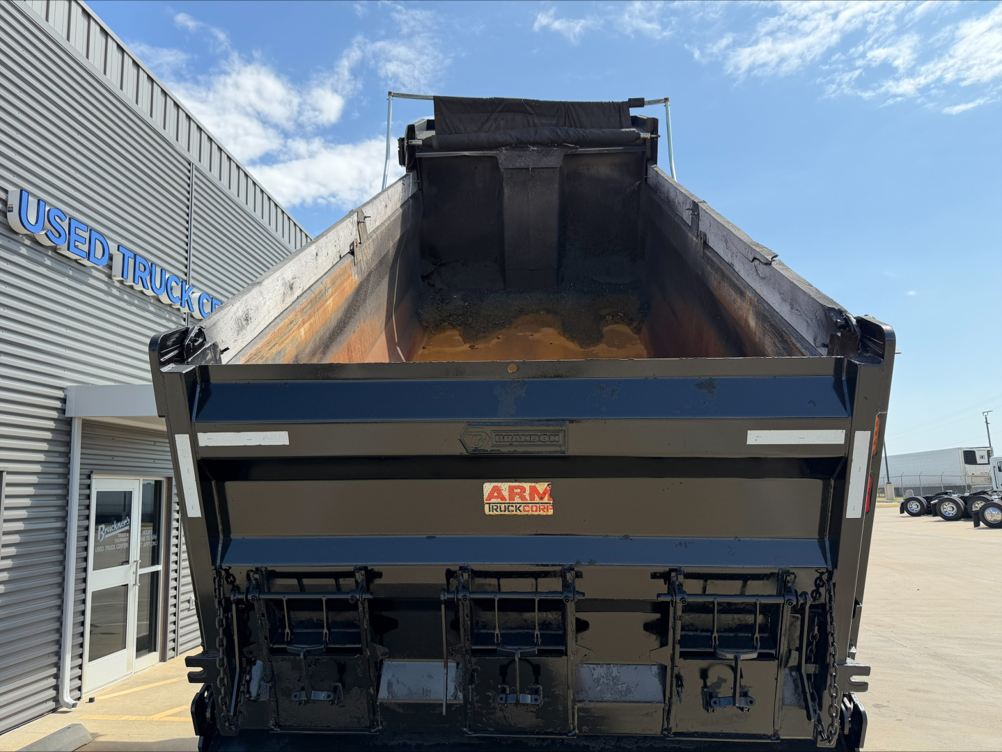 Dump truck bed of a 2023 Mack GR84F with open-box and tailgate, showing metal sides and cargo area; arm hydraulics visible; dealership building in background.
