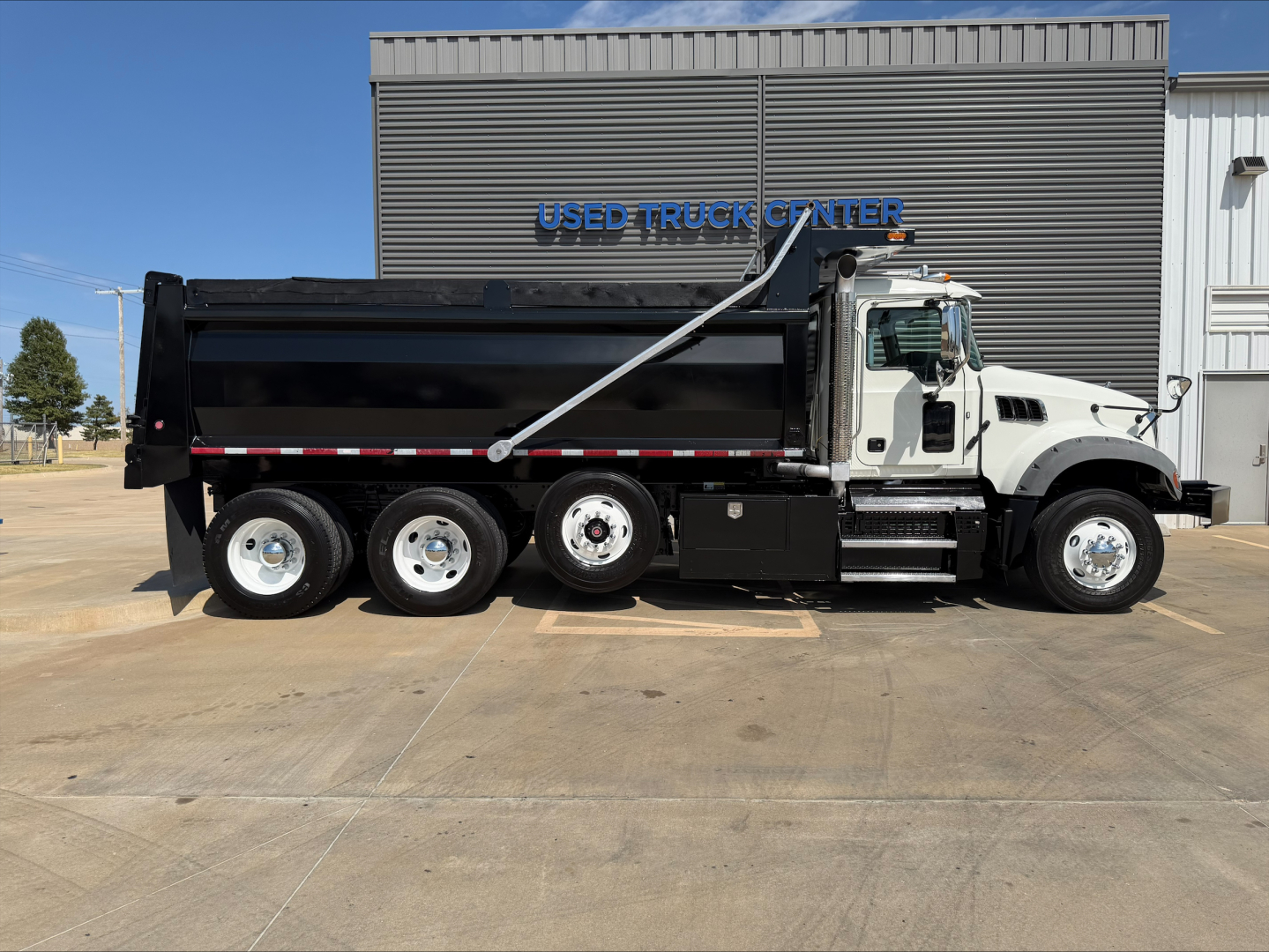 Truck side profile showing a white 2023 Mack GR84F dump truck parked in front of a building with blue 'USED TRUCK CENTER' sign. The dump bed is black with a silver hydraulic arm and chrome wheels.