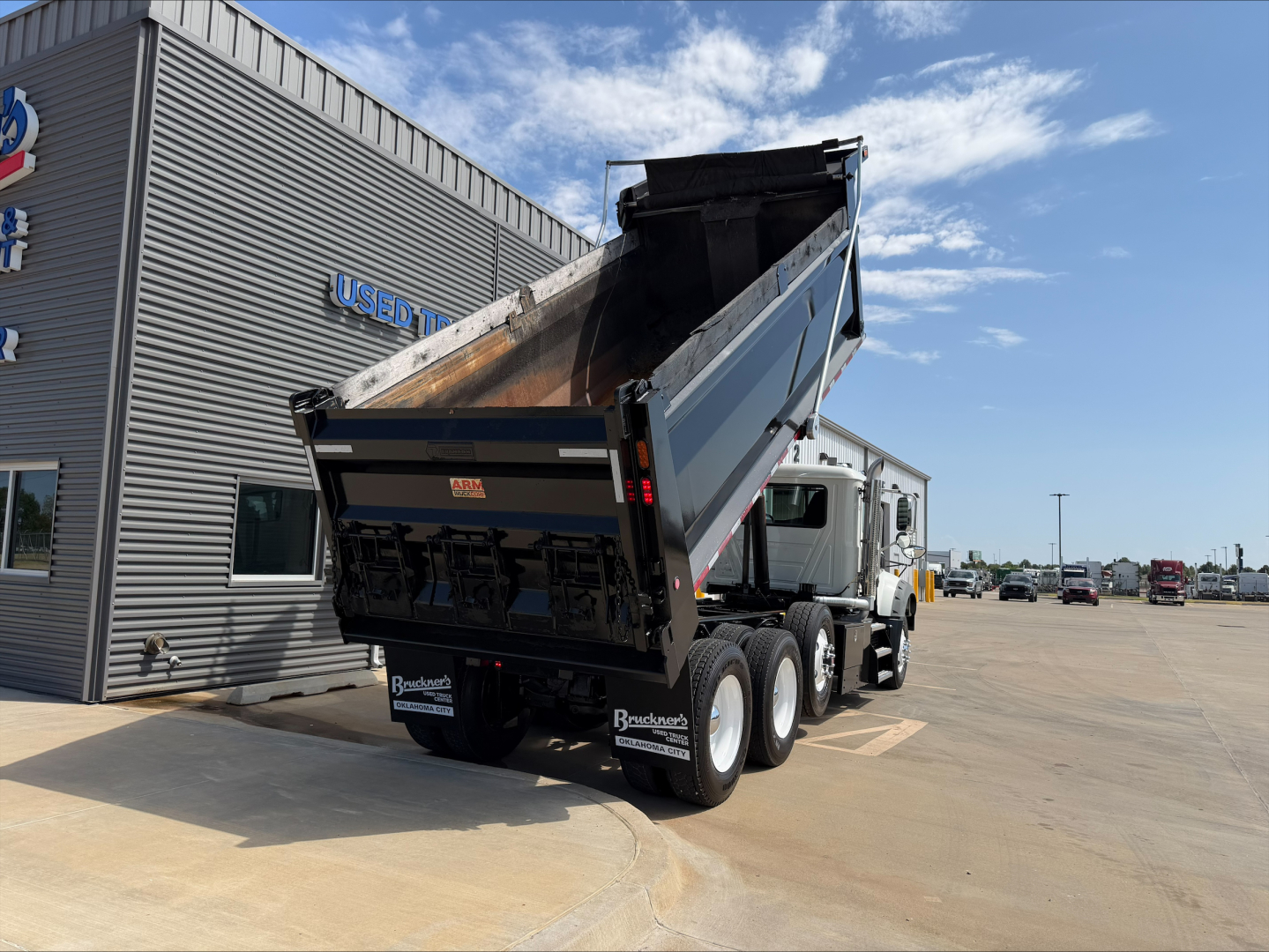 Dump truck in a dealership lot with raised bed, Mack GR84F model, chrome wheels, and white cab; trailer hitch visible; stock number plate and dealership building in background.
