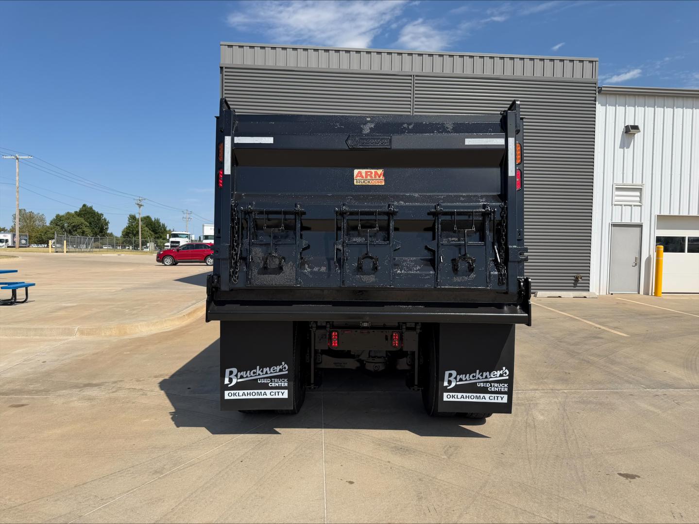 Rear view of a 2023 Mack GR84F dump truck with a black dump body and dual rear mudflaps that read Brauckers Oklahoma City; visible white logos on the black tailgate; clear blue sky.
