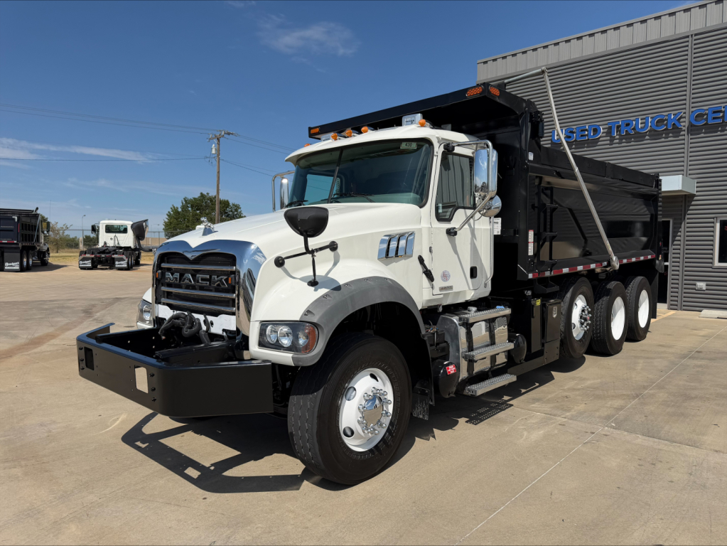 Mack GR84F dump truck in a dealership lot with white cab and dump bed, front bumper guard, large grille, dual rear axles, and side step. Visible text includes MACk on grille and truck body details.