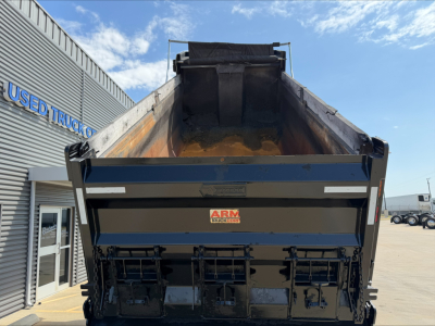 Dump truck bed of a 2023 Mack GR84F with open-box and tailgate, showing metal sides and cargo area; arm hydraulics visible; dealership building in background.