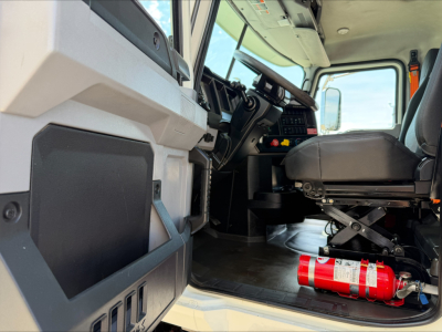 Truck cab interior of a 2023 Mack GR84F showing driver seat, dashboard, and a red fire extinguisher under the seat.