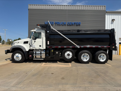 Dump truck side view showing 3-axle Mack GR84F with white cab and black dump body, in front of a blue sign building.