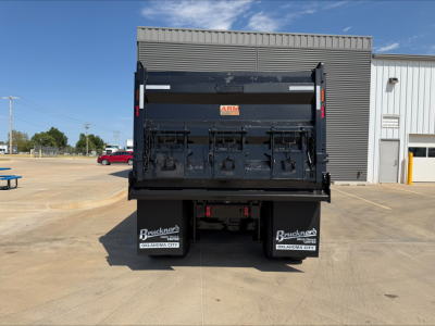 Rear view of a 2023 Mack GR84F dump truck with a black dump body and dual rear mudflaps that read Brauckers Oklahoma City; visible white logos on the black tailgate; clear blue sky.