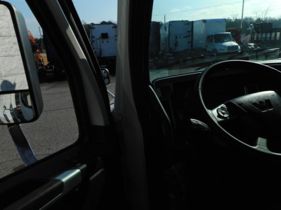 Truck cab interior with steering wheel and dashboard, side window view of a lot with other trucks and vehicles
