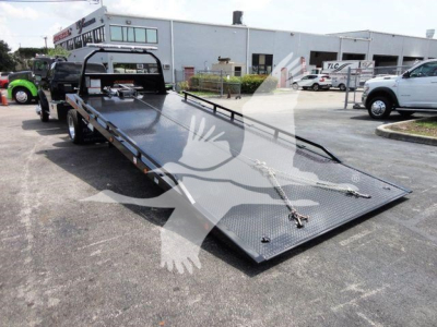 Ram 5500 rollback tow truck flatbed with a lowered black steel bed and ramp in a dealership lot, rear view showing long flatbed and side rails.