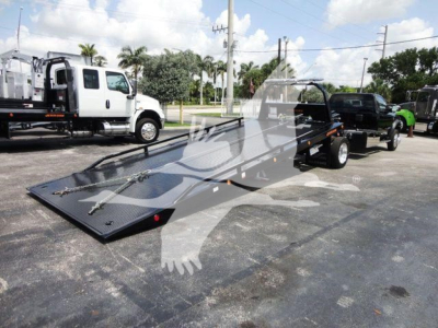 Ram 5500 rollback tow truck with a long low-profile flatbed and a black metallic finish, dual rear wheels, and a white cab in background.