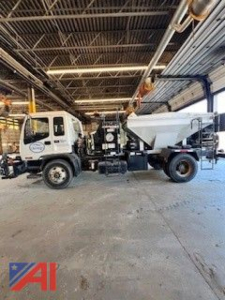 GMC T7500 pothole patcher truck in an indoor shop, side view showing cab, chassis, and large hopper with piping overhead.