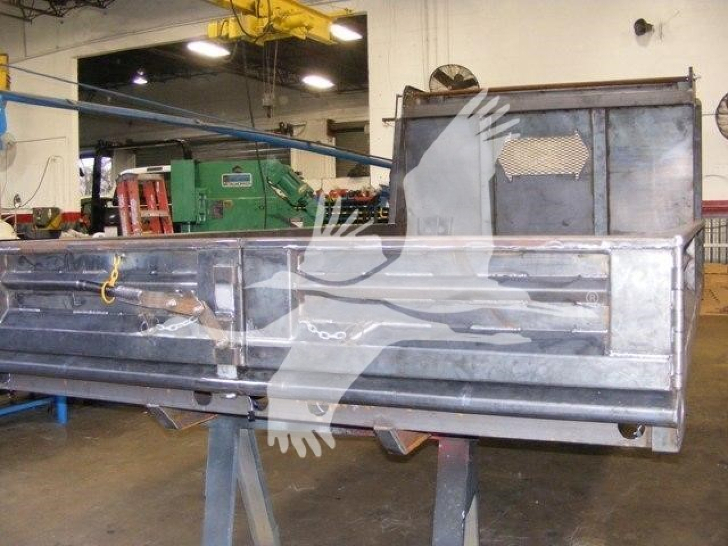 Dump truck bed on a metal frame showing a large silver cargo box with a cutout rear panel and a windowed cab area behind. Industrial workshop with green machinery in background.