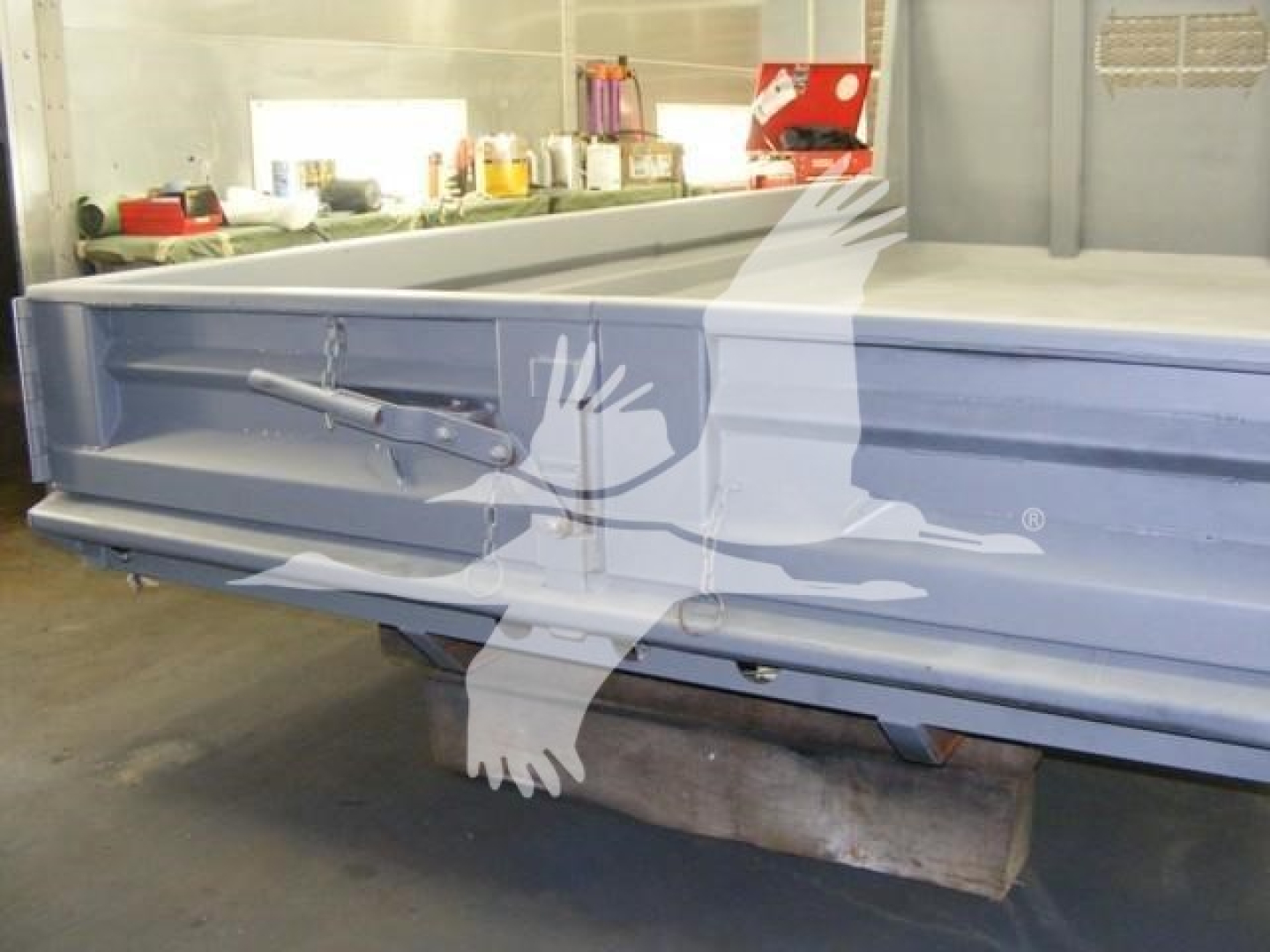Dump truck bed showing side-mounted tailgate latch and hydraulic rod in a workshop.