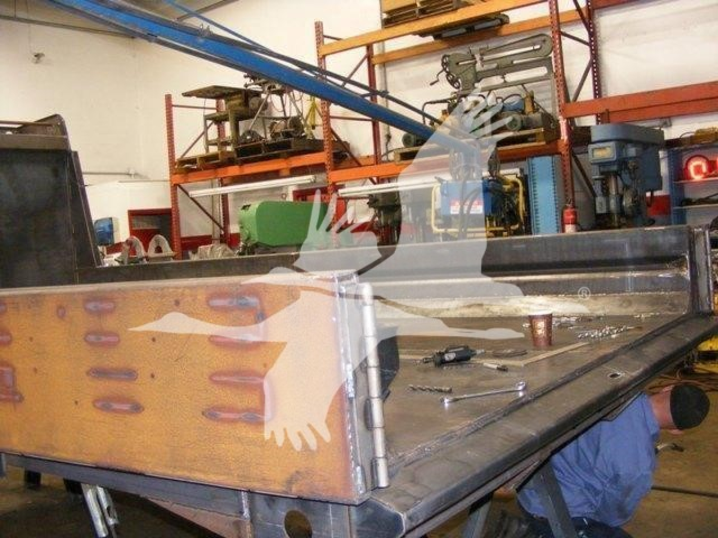 Dump truck bed or tailgate component with metal panels and wooden side accent, inside workshop with equipment and shelving in background.