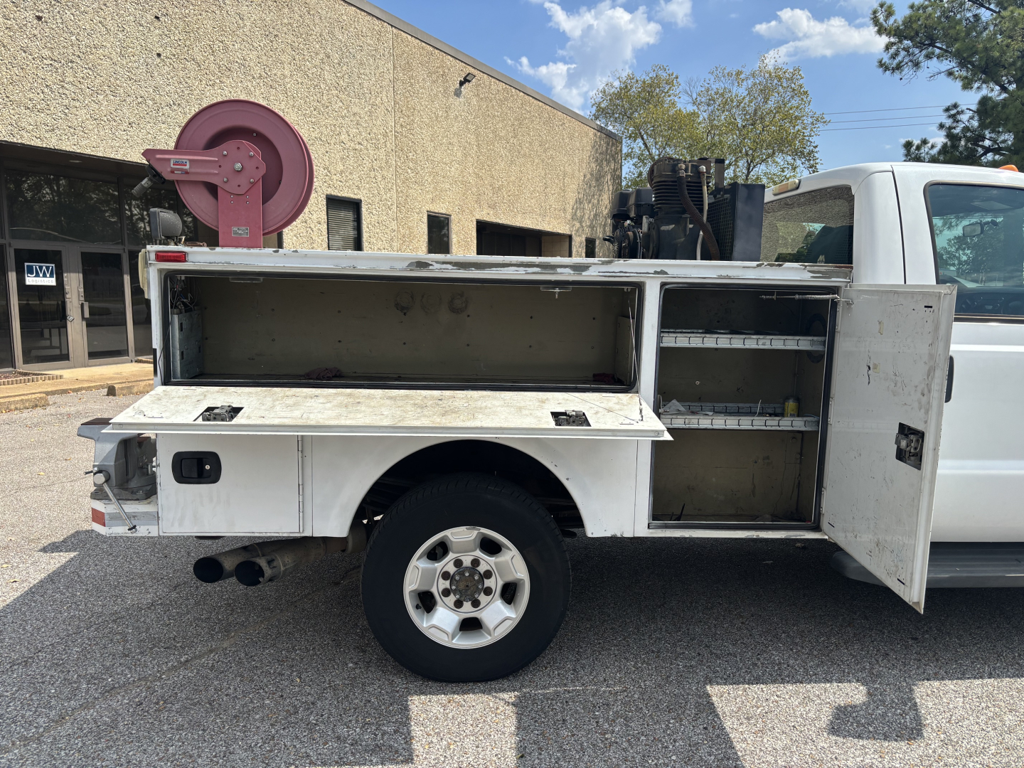 Side view of a white Ford service utility truck with external utility/storage compartments open, showing tool trays and storage shelves in the bed. Dual rear exhaust visible.