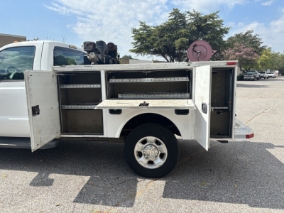 Ford F-250 utility service body side view showing multiple storage compartments and open cabinet doors, white cargo bed with toolbox, side compartments, and wheel with hubcap.