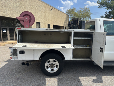 Side view of a white Ford service utility truck with external utility/storage compartments open, showing tool trays and storage shelves in the bed. Dual rear exhaust visible.