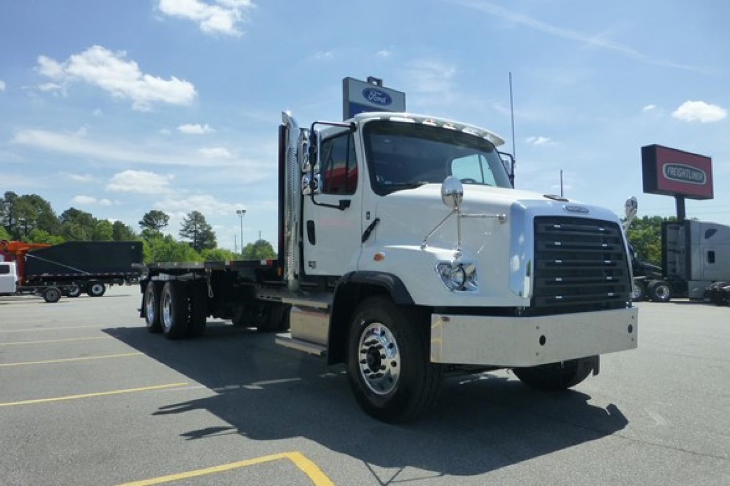 Freightliner 114SD Plus flatbed truck in white, side view, daytime with dealership lot in background.