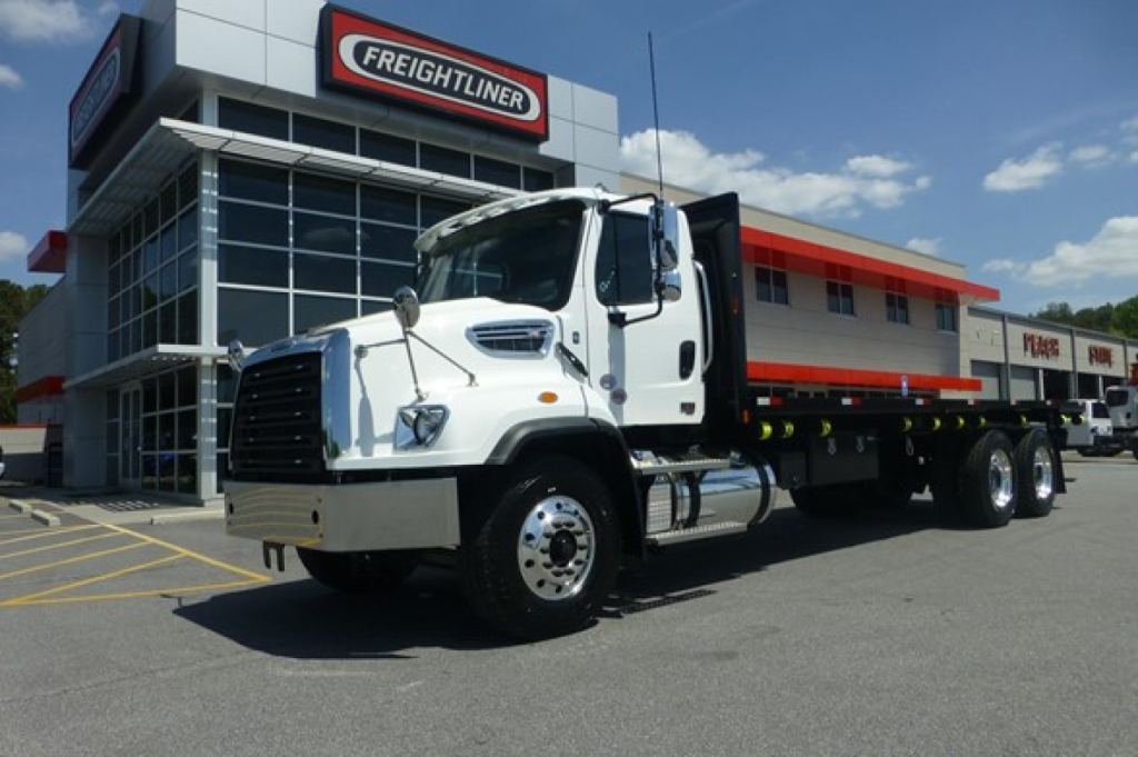 Freightliner 114SD Plus flatbed truck in front of dealership showing cab and long flatbed trailer.