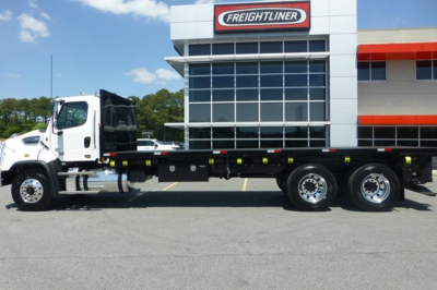  Freightliner 114SD Plus flatbed truck side view in front of dealership, showing cab, flatbed with side rails, tandem rear axles, and Freightliner branding on building.