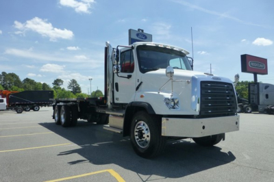 Freightliner 114SD Plus flatbed truck in white, side view, daytime with dealership lot in background.