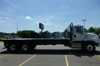 Flatbed truck in a dealership lot showing a Freightliner 114SD Plus chassis with a long flatbed and cab, side view.