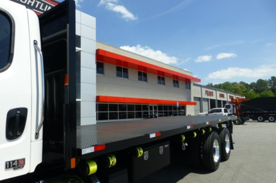 Freightliner 114SD Plus flatbed truck with long steel deck and dual rear axles, white cab, flatbed in view, dealership lot in background.