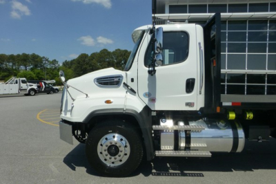 Freightliner 114SD Plus truck cab side view showing front wheel, chrome fuel tank, side steps, and dump bed truck in a dealership lot.