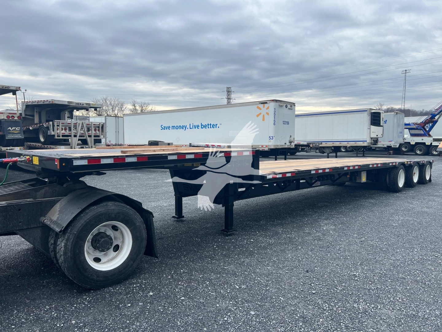 Trailer side view showing a drop deck middle lift with two axles, white cabover trailers in background, long flatbed with black frame and white wheels.
