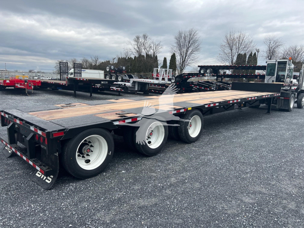 Tri-axle drop deck trailer with wood deck and three axles visible, rear-ratio wheels, black frame, white rims, reflective tape, overall in-service industrial setting.