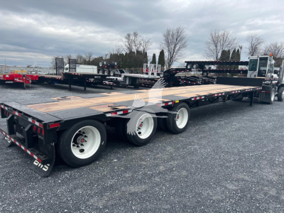 Tri-axle drop deck trailer with wood deck and three axles visible, rear-ratio wheels, black frame, white rims, reflective tape, overall in-service industrial setting.