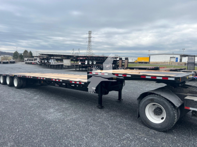 Flatbed trailer with drop middle lift and multiple axles in outdoor lot, black deck, white wheels, equipment in background.