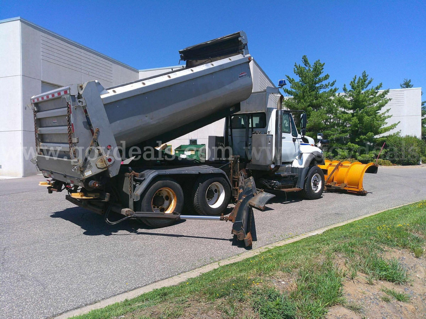 Dump truck with snow plow attachment and dump bed raised, front-right view showing yellow plow blade and debris bucket on ground.