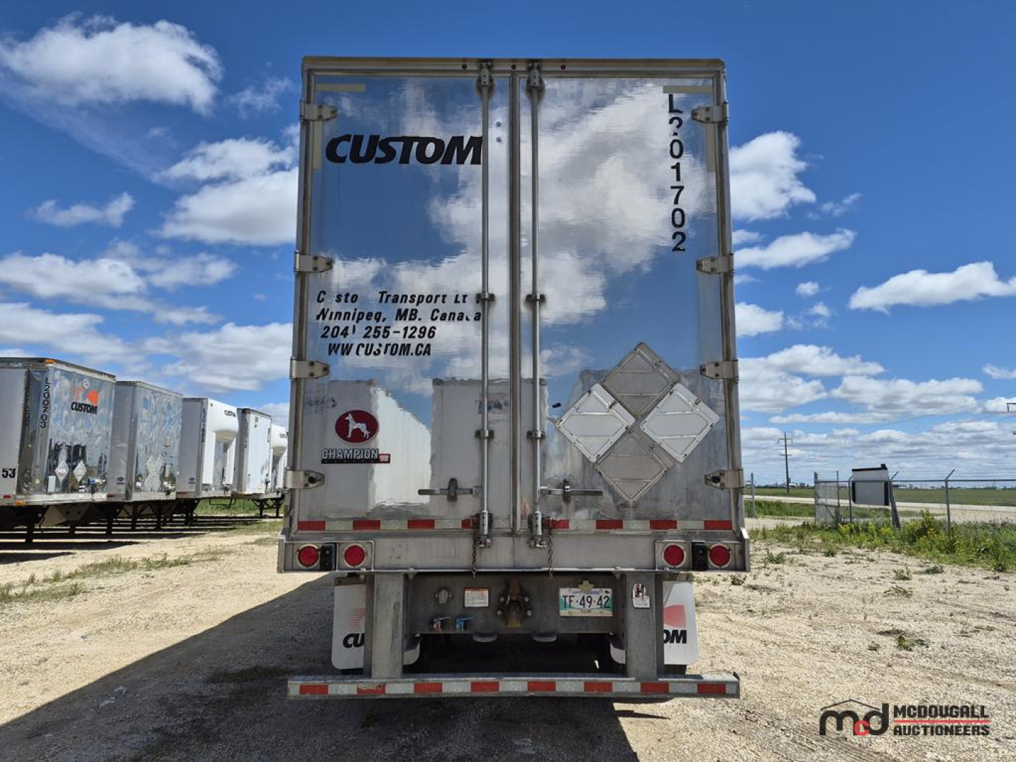 Trailer rear showing Custom logo on doors, reflective diamonds, tail lights, and bumper; vertical VIN/sticker along right door edge; clouded sky with other trailers in lot.