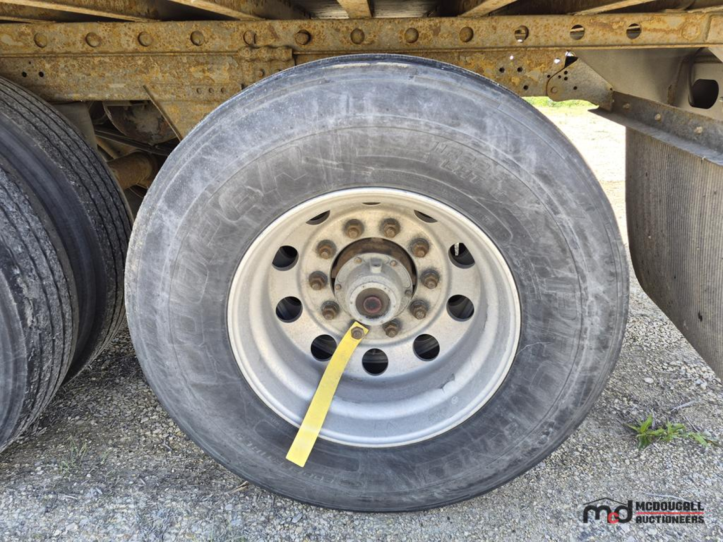 Trailer wheel close-up showing 53' dry van semi trailer with silver steel wheel, black tire, yellow strap tied around lug nut, rusty frame in background.