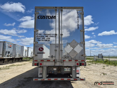 Trailer rear showing Custom logo on doors, reflective diamonds, tail lights, and bumper; vertical VIN/sticker along right door edge; clouded sky with other trailers in lot.