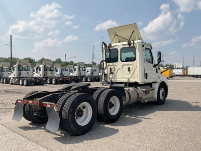 Freightliner Cascadia126 tractor chassis with 3 axles, white cab, exposed fifth wheel and dual rear tires, raised sleeper, in yard.