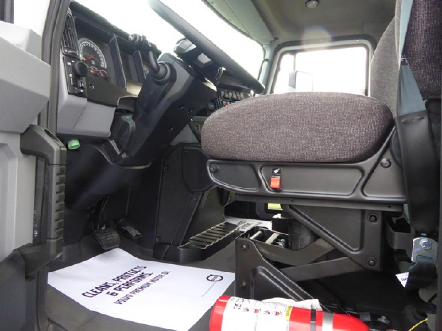 Interior cab view of a 2025 Mack MD6 truck showing driver’s seat, steering wheel, and dashboard with pedals; floor mat with text; part of passenger area.