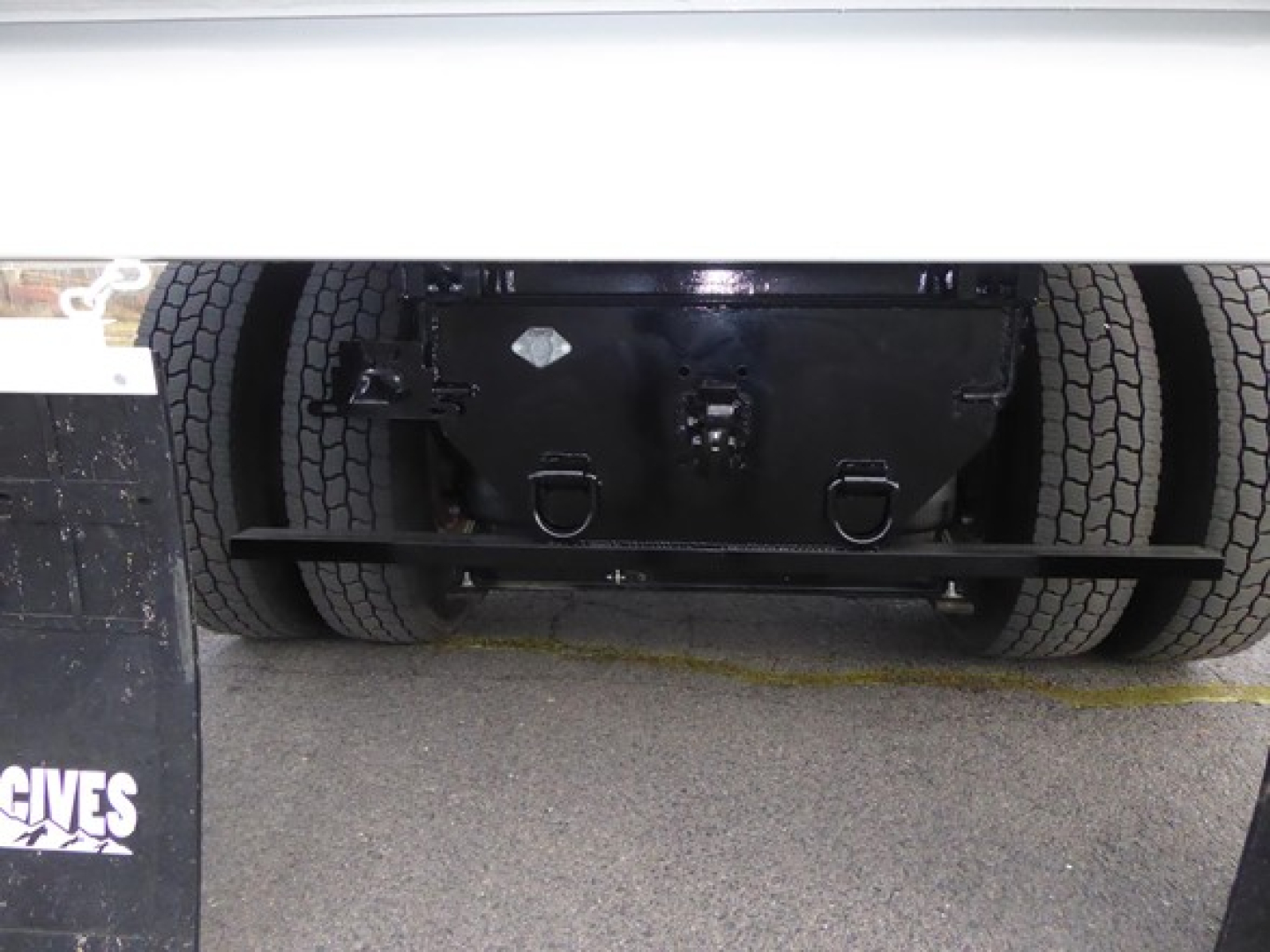 Front underside of a 2025 Mack MD6 dump truck showing rear axle and frame with dual tires and black protective skid, visible undercarriage components.
